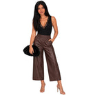 A woman modeling a pair of high-waisted, wide-leg brown faux leather pants with a black lace-trimmed sleeveless top, holding a black clutch against a plain white background.