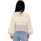 A woman viewed from the back wearing a cream-colored sheer lace blouse with ruffled details and long bell sleeves, paired with light blue jeans, against a plain white background.