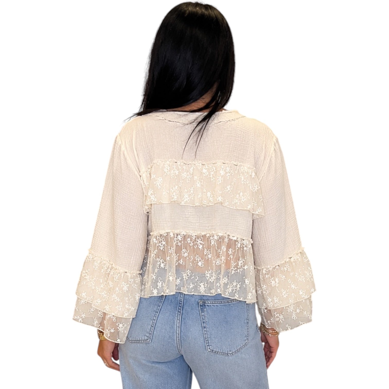 A woman viewed from the back wearing a cream-colored sheer lace blouse with ruffled details and long bell sleeves, paired with light blue jeans, against a plain white background.