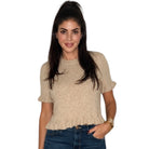 A woman wearing a beige short-sleeve knit top with ruffled cuffs and a ruffled hem, paired with blue jeans, against a plain white background.