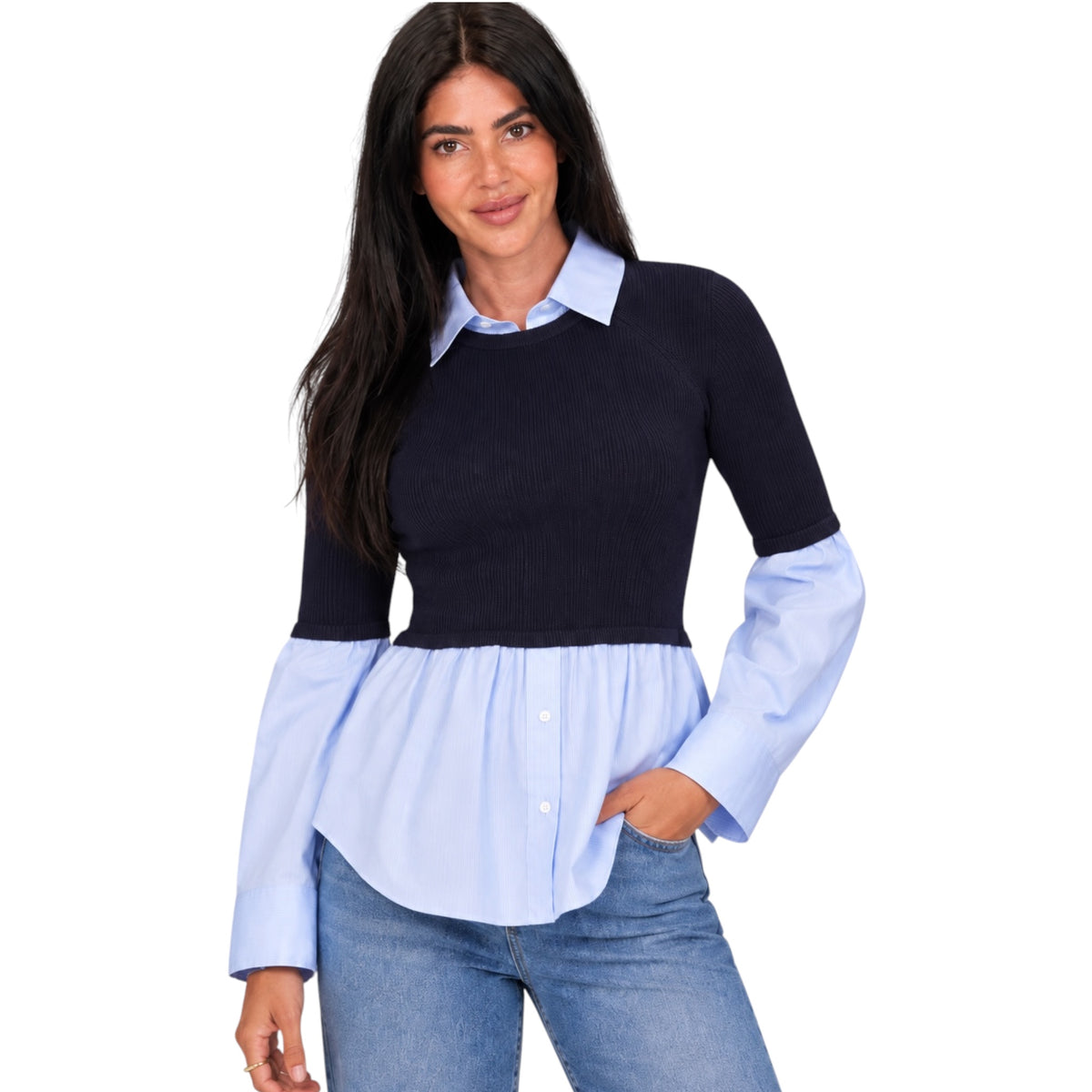A woman modeling a long-sleeve knit top with a dark navy ribbed upper section and light blue button-up shirt sleeves and collar, paired with blue jeans against a plain white background.