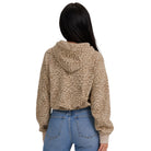 A woman viewed from the back wearing a beige and brown leopard-print cropped hoodie with light beige ribbed cuffs, paired with blue jeans, against a plain white background.