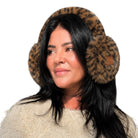 A woman wearing plush leopard-print earmuffs with a small tan brand patch on the right earcup, paired with a cream knit sweater against a plain white background.