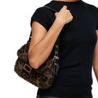 A person wearing a black short-sleeve top carries a brown and black leopard-print faux fur shoulder bag with gold-tone hardware against a plain white background.