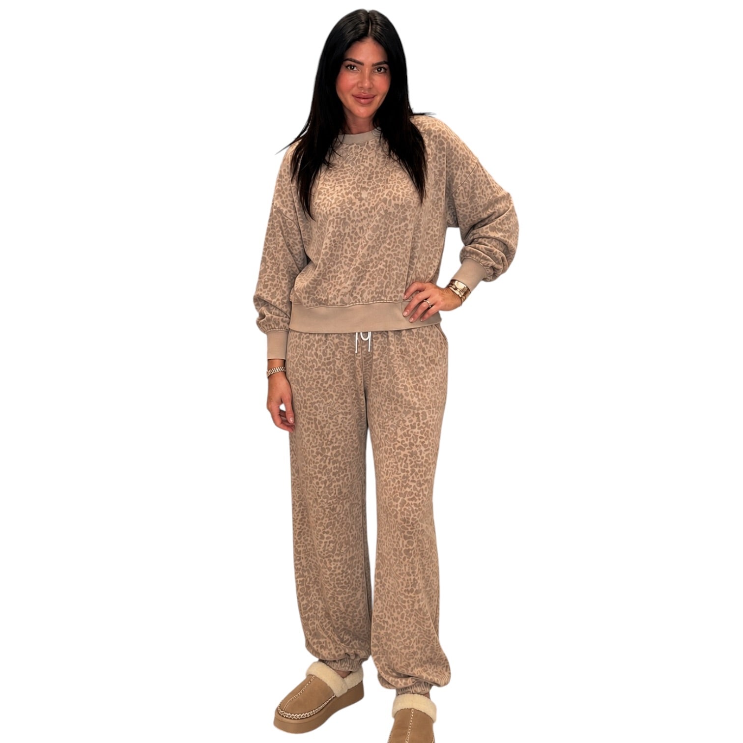 A woman modeling a matching beige and brown leopard-print lounge set, consisting of a long-sleeve sweatshirt and jogger pants, paired with tan slippers, against a plain white background.