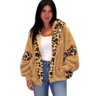 A woman modeling a beige sherpa zip-up hoodie with leopard print panels on the hood, cuffs, and sleeves, worn over a white tank top and blue jeans against a plain white background.