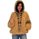 A woman wearing a tan sherpa zip-up hoodie with a leopard print hood, zipper placket, and sleeve patches, paired with blue jeans, against a plain white background.
