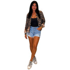 A woman modeling a leopard print jacket over a black tank top, paired with light blue denim shorts and white sneakers, set against a plain white background.
