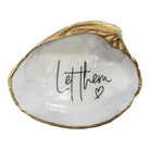 A decorative clam shell-shaped tray with a textured gold-colored rim and a glossy white interior featuring the black handwritten script 'Let them' and a small heart.