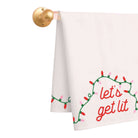 A white kitchen towel hanging from a gold wall-mounted rod, printed with a green Christmas light garland and the phrase 'let's get lit' in red script.