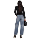 A woman shown from the back wearing light-wash blue wide-leg jeans with large patch pockets, a black long-sleeve top, and black high heels, while holding a black woven handbag against a plain white background.