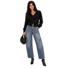 A woman modeling a black button-front cardigan, wide-leg blue denim jeans with patch pockets, black ankle boots, and holding a black woven handbag against a plain white background.