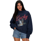 A woman modeling a navy blue sweatshirt with a red 'Lucky' script and a flying mallard duck graphic on the front, paired with denim shorts against a plain white background.
