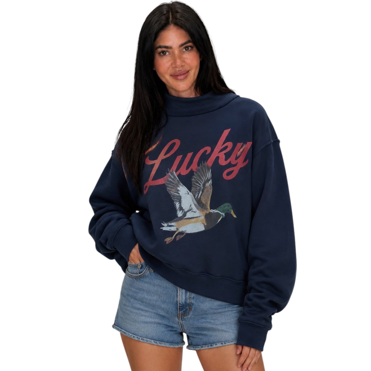 A woman modeling a navy blue sweatshirt with a red 'Lucky' script and a flying mallard duck graphic on the front, paired with denim shorts against a plain white background.