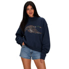 A woman modeling a navy blue sweatshirt with a detailed brown and green duck graphic on the front, paired with denim shorts against a plain white background.
