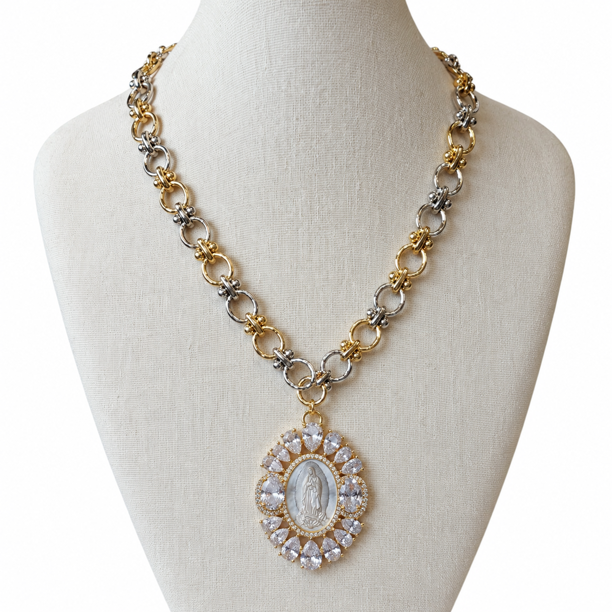 A two-tone gold and silver chain necklace with a circular pendant featuring a white religious image surrounded by clear rhinestones, displayed on a cream fabric mannequin bust.