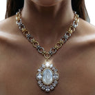 A close-up of a woman's neck and chest wearing a multi-metal necklace with a large oval religious pendant featuring a white figure surrounded by clear stones.