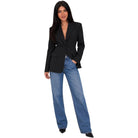 A woman modeling a black blazer with wide-leg blue jeans and black pointed shoes, standing against a plain white background.