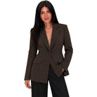 A woman modeling a dark brown single-breasted blazer with notch lapels and flap pockets, paired with black trousers, against a plain white background.