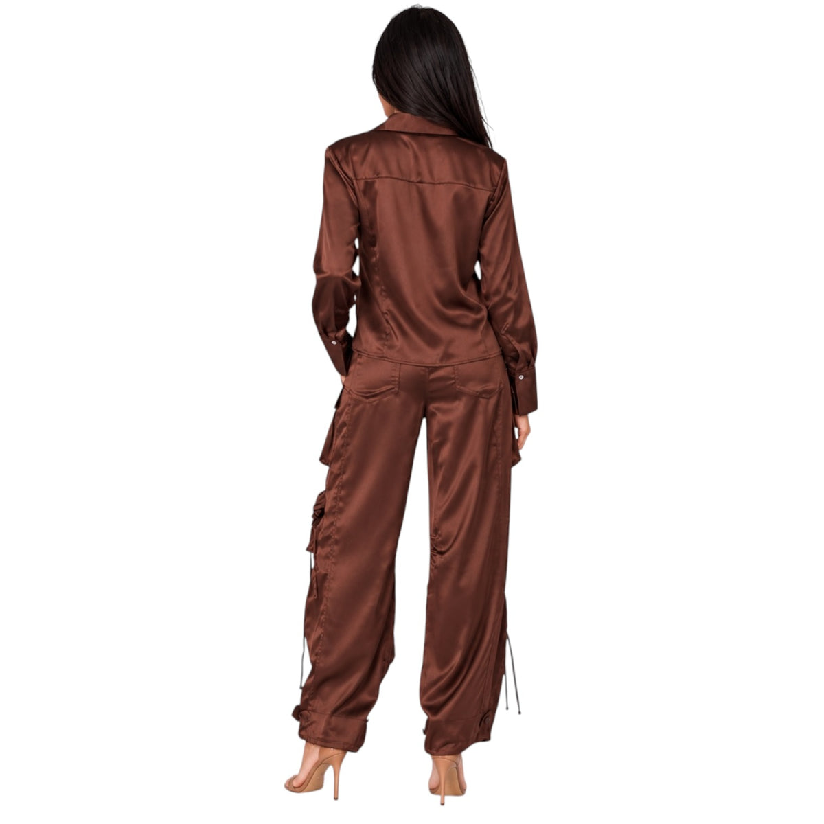 A woman viewed from the back wearing a shiny brown satin jumpsuit with side zipper details, paired with nude high heels, against a plain white background.