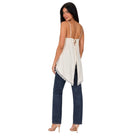 A woman shown from the back wearing a cream-colored spaghetti-strap top with lace trim and a tie-back detail, paired with dark blue jeans and nude heels, against a plain white background.