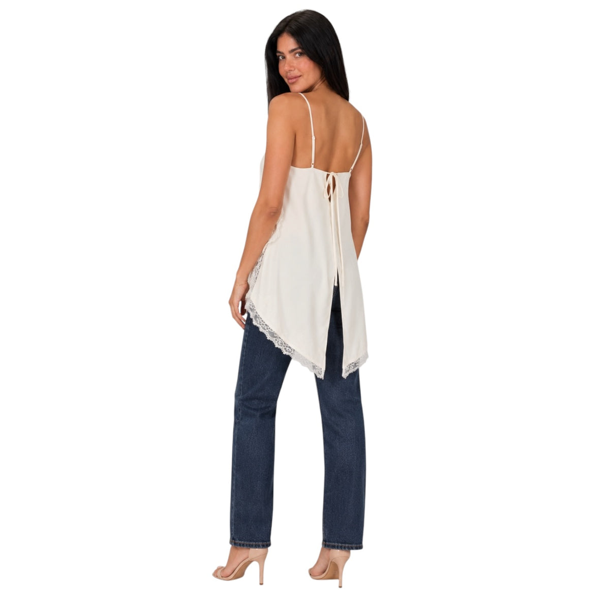 A woman shown from the back wearing a cream-colored spaghetti-strap top with lace trim and a tie-back detail, paired with dark blue jeans and nude heels, against a plain white background.