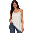 A woman modeling a cream-colored camisole top with delicate lace trim along the edges and neckline, paired with blue jeans against a plain white background.