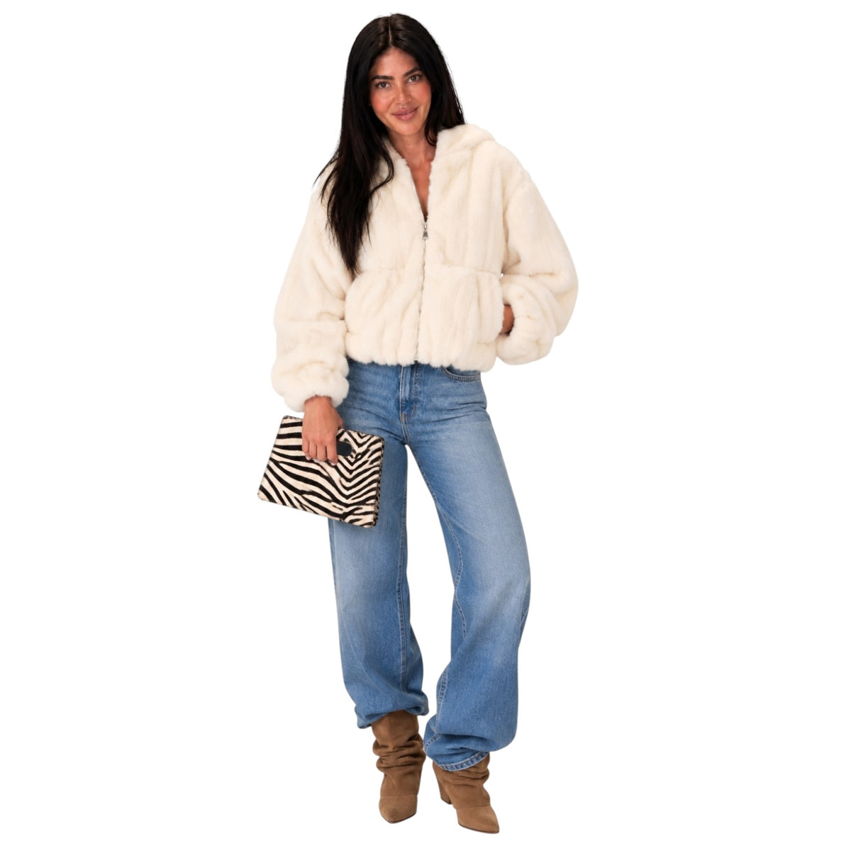 A woman modeling a cream-colored faux fur zip-up jacket, paired with blue jeans, brown boots, and holding a black and white zebra print clutch, set against a plain white background.