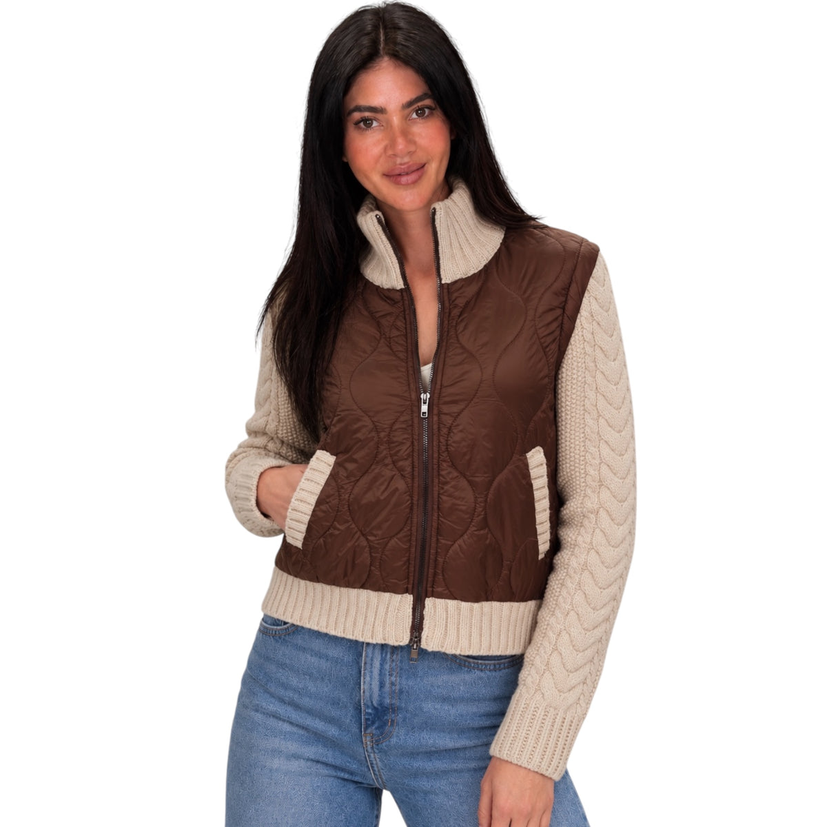 A woman modeling a brown quilted zip-front vest with cream cable-knit sleeves, ribbed collar and hem, paired with blue jeans against a plain white background.