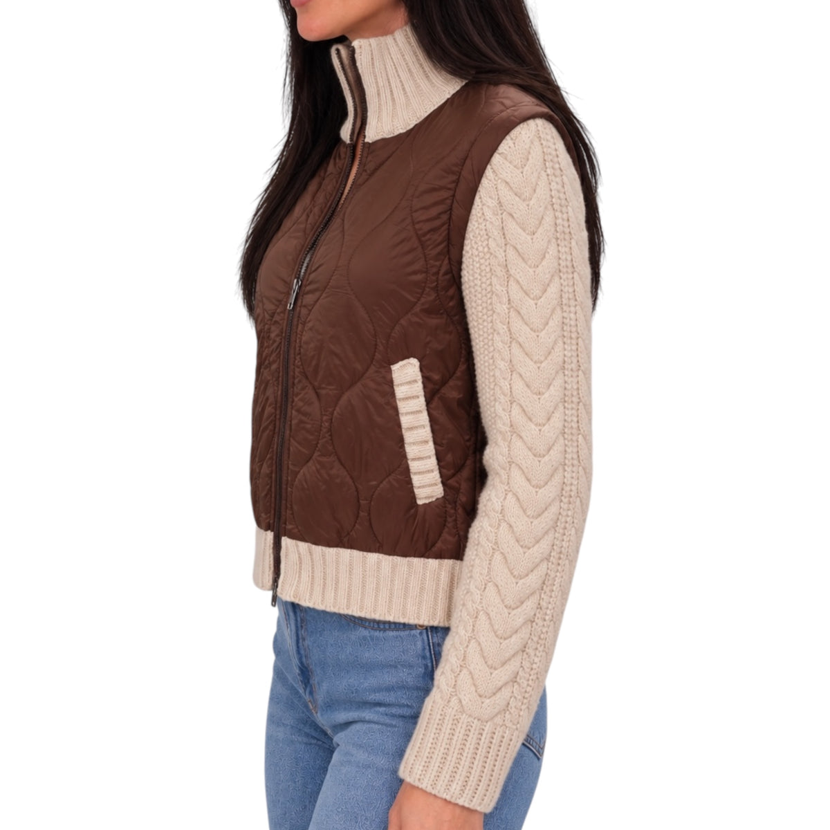 A woman wearing a brown quilted sleeveless zip-front vest over a cream cable-knit mock-neck sweater and blue jeans, shown against a plain white background.