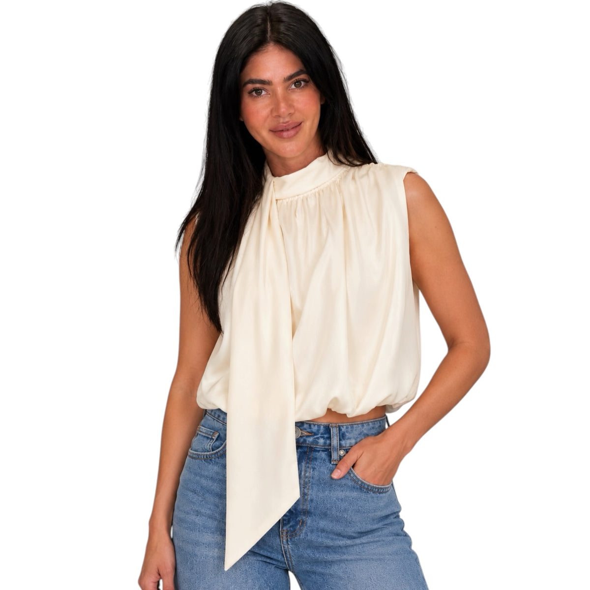 A woman modeling a sleeveless cream blouse with a high gathered neckline and a long draped tie detail, paired with blue jeans against a plain white background.