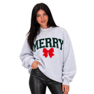 A woman modeling a light heather gray crewneck sweatshirt with 'MERRY' in dark green varsity letters and a red bow graphic on the front, paired with black faux-leather pants against a plain white background.