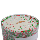 A close-up of a holiday-themed candle in a clear glass container, filled with white wax and decorated with red and green sprinkles around the rim and on the surface. A small piece of natural wood is centered as the wick.