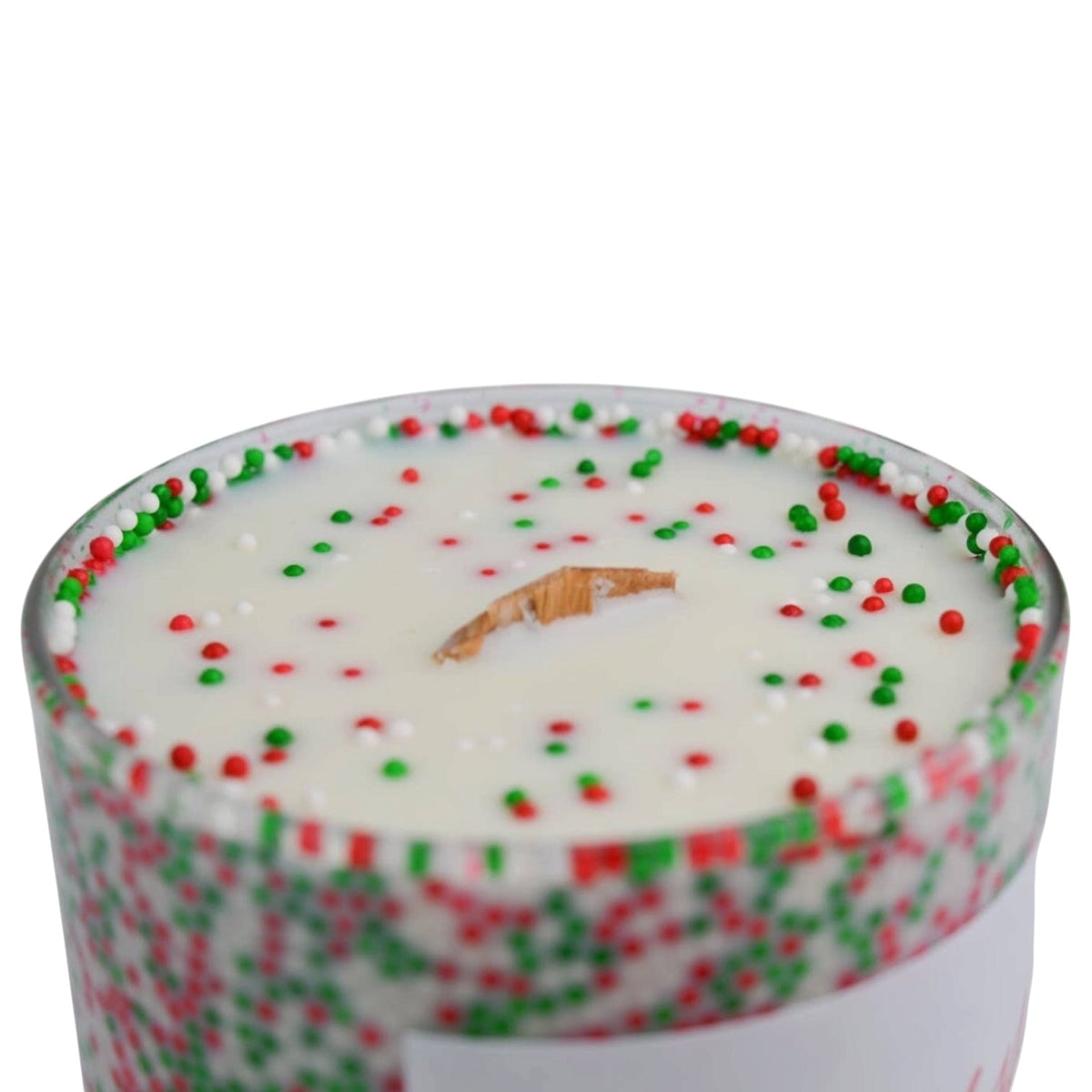 A close-up of a holiday-themed candle in a clear glass container, filled with white wax and decorated with red and green sprinkles around the rim and on the surface. A small piece of natural wood is centered as the wick.