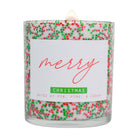 A lit scented candle in a clear glass jar filled with white wax and decorated with red and green holiday sprinkles. The front label reads 'merry CHRISTMAS' with 'NOTES OF FIR, PINE, & CEDAR' below, on a white background.