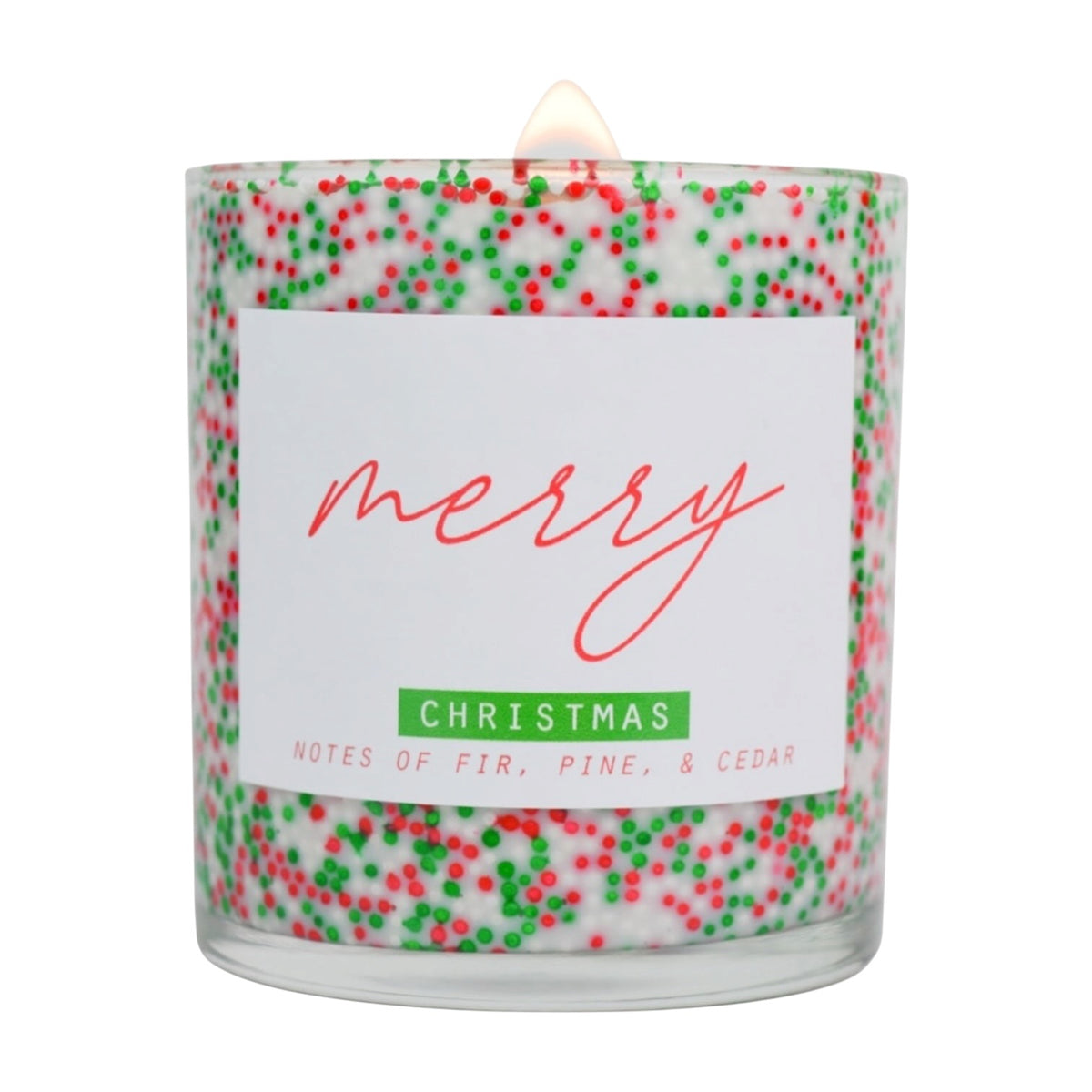 A lit scented candle in a clear glass jar filled with white wax and decorated with red and green holiday sprinkles. The front label reads 'merry CHRISTMAS' with 'NOTES OF FIR, PINE, & CEDAR' below, on a white background.