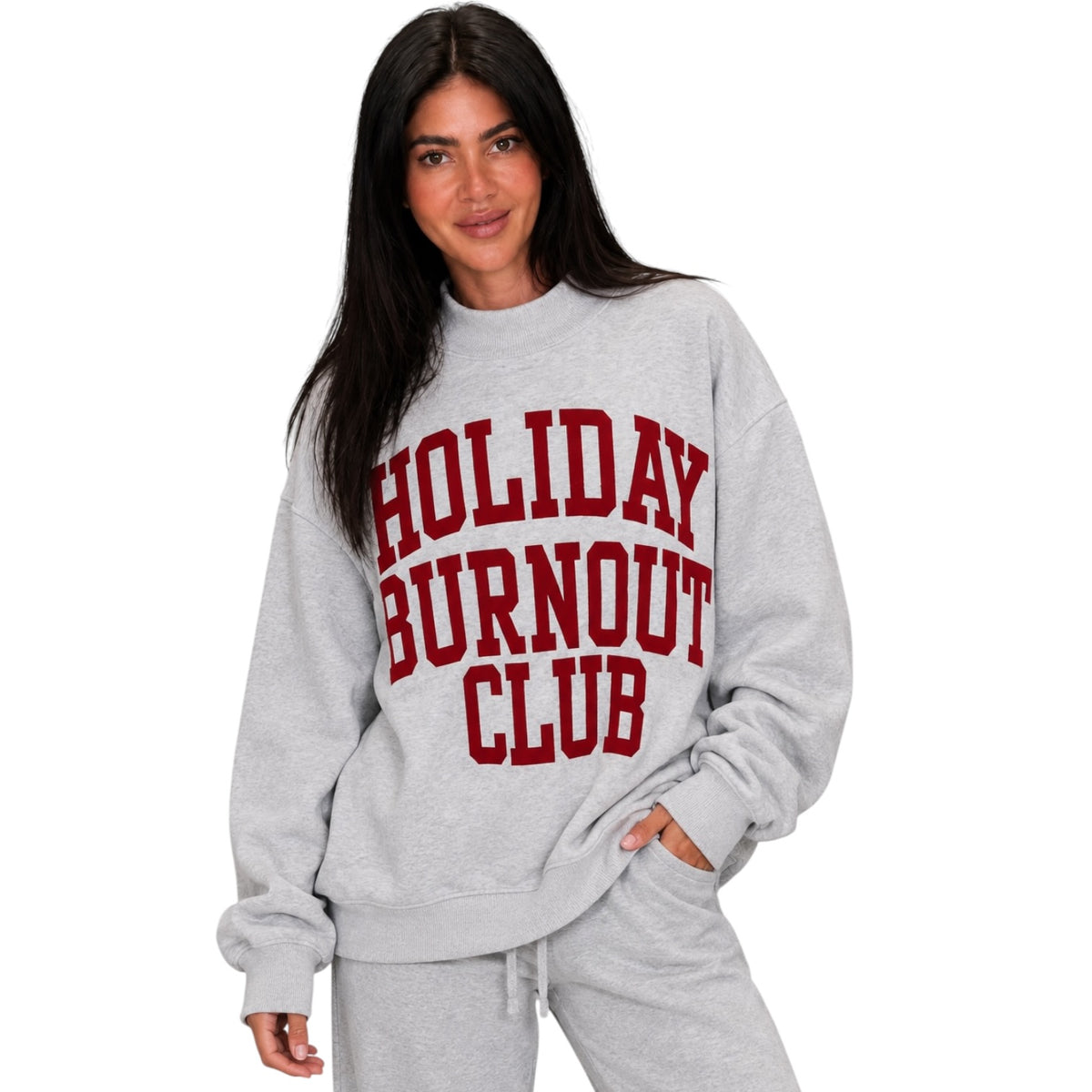 A woman modeling a light heather gray crewneck sweatshirt with 'HOLIDAY BURNOUT CLUB' printed in bold red varsity letters across the chest, paired with matching gray sweatpants against a plain white background.