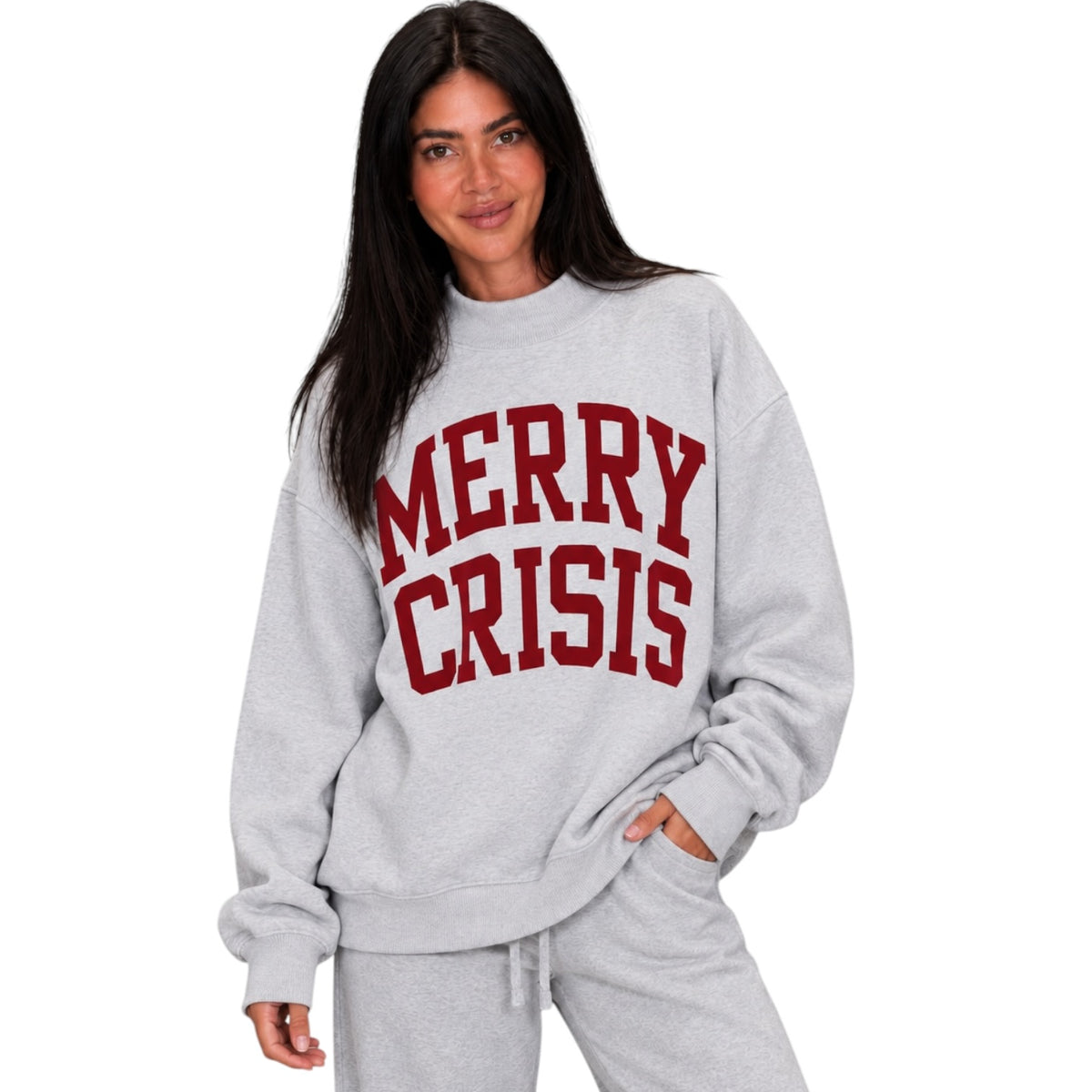 A woman modeling a light heather gray sweatshirt with 'MERRY CRISIS' printed in red varsity-style letters across the chest, paired with matching gray sweatpants against a plain white background.