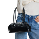 A black leather duffel-style handbag with two top handles, carried over the shoulder of a person wearing a cream draped top and blue jeans against a plain white background.
