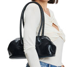 A woman wearing a cream-colored top and blue jeans carries a glossy black leather shoulder bag against her shoulder, shown from the side against a plain white background.
