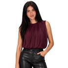 A woman modeling a sleeveless pleated top in deep burgundy, paired with black faux-leather pants, against a solid white background.