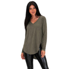 A woman modeling a long-sleeve, V-neck, high-low tunic top in olive green, paired with black faux-leather leggings, against a plain white background.