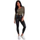 A woman modeling a long-sleeve olive green crop top and shiny black high-waisted leggings with white sneakers, set against a plain white background.