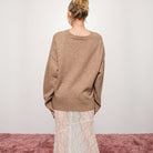 Person wearing a mocha sweater and light-colored pants standing on a pink surface with a white background