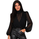 A woman modeling a black sheer long-sleeve blouse with a high mock neck and a wrap-style front overlay, paired with black faux-leather pants, against a plain white background.