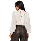 A woman shown from the back wearing a cream-colored long-sleeve blouse with a large bow detail at the back neckline, paired with dark brown faux-leather pants, against a plain white background.