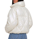 A model shown from the back wearing a shiny, puffer-style jacket with a high collar and quilted diamond pattern, paired with blue jeans against a plain white background.