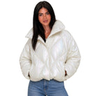 A woman modeling a shiny, puffer-style cropped jacket in a metallic cream or off-white color, paired with blue jeans, against a plain white background.