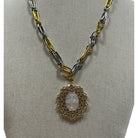 A two-tone gold and silver chain necklace with a circular pendant featuring a central white oval stone surrounded by small clear stones, displayed on a beige fabric jewelry bust.