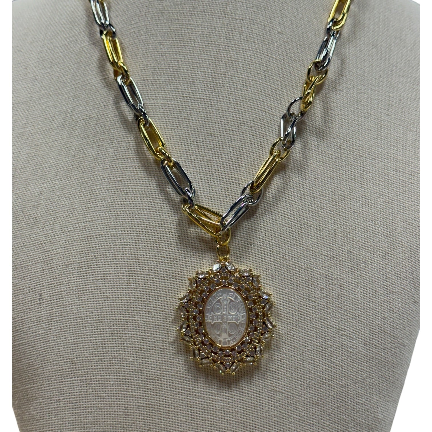 A two-tone gold and silver chain necklace with a circular pendant featuring a central white oval stone surrounded by small clear stones, displayed on a beige fabric jewelry bust.